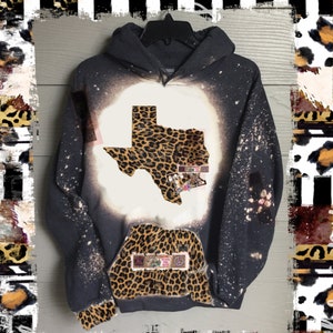 May include: A black hoodie with a bleach-splattered design. The hoodie features a white outline of the state of Texas filled with a leopard print pattern. The pocket of the hoodie also features a leopard print pattern with a small patch that says "Peace Love Music".