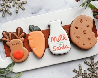 May include: A set of four Christmas-themed cookies on a white plate. The cookies are decorated with icing and include a reindeer, a carrot, a bottle of milk with the text "Milk for Santa", and a chocolate chip cookie.