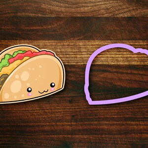 May include: A cartoon taco cookie cutter with a smiling face. The cutter is made of purple plastic and is shaped like a taco. The cutter is on a wooden surface.