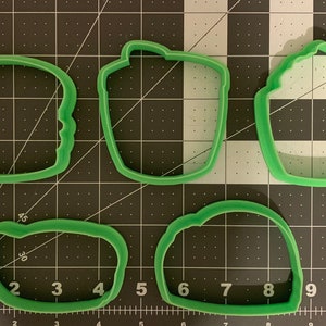 May include: Four green plastic cookie cutters shaped like a hamburger, a milkshake, a french fry container, and a hot dog. The cutters are on a black and white grid background.