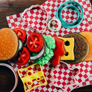 Puede incluir: Un juego de cortadores de galletas con forma de hamburguesa, queso, lechuga, tomate y cebolla. Los cortadores est&aacute;n sobre un mantel a cuadros rojos y blancos.