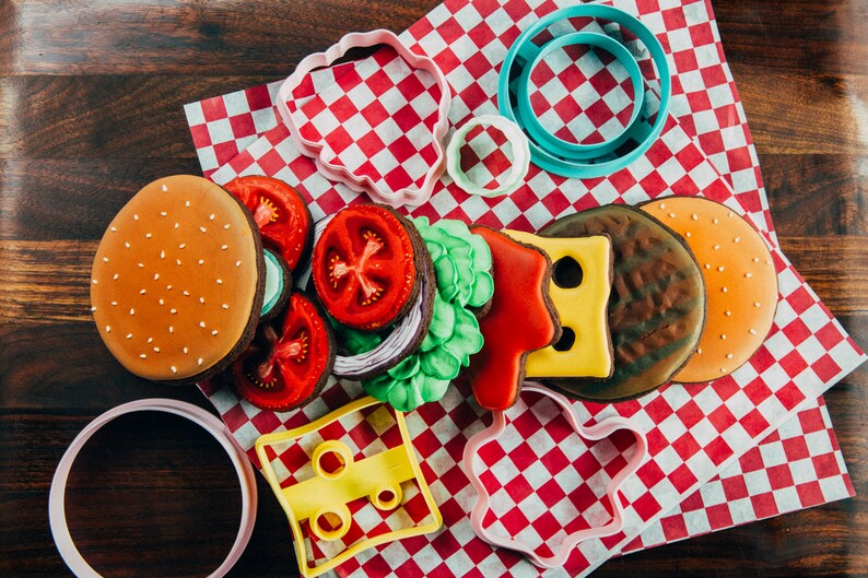 Puede incluir: Un juego de cortadores de galletas con forma de hamburguesa, queso, lechuga, tomate y cebolla. Los cortadores est&aacute;n sobre un mantel a cuadros rojos y blancos.