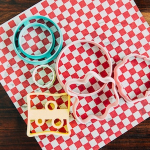 Puede incluir: Un conjunto de seis cortadores de galletas de colores sobre un fondo a cuadros rojo y blanco. Los cortadores tienen forma de c&iacute;rculo, nube, estrella, rect&aacute;ngulo y dos c&iacute;rculos de diferentes tama&ntilde;os.