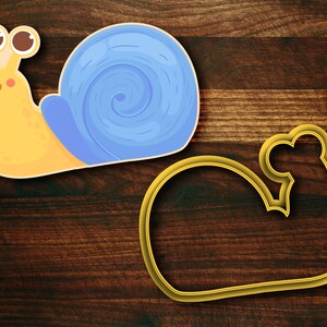 May include: A yellow snail cookie cutter with a blue shell and a smiling face. The cutter is on a wooden surface next to a gold-coloured outline of the snail.