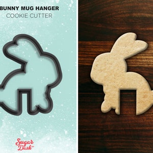 May include: A black cookie cutter in the shape of a bunny rabbit with a cutout for a mug handle. The bunny is standing on its hind legs with its ears up. The cookie cutter is on a light blue background with white speckles. The text "BUNNY MUG HANGER COOKIE CUTTER" is above the cookie cutter. The text "Sugar Dash" is below the cookie cutter.