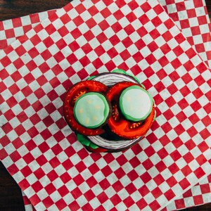 Puede incluir: Un papel a cuadros rojo y blanco con una galleta en capas decorada para parecer una hamburguesa. La hamburguesa de galleta tiene glaseado rojo y verde y un c&iacute;rculo de glaseado blanco en la parte superior.