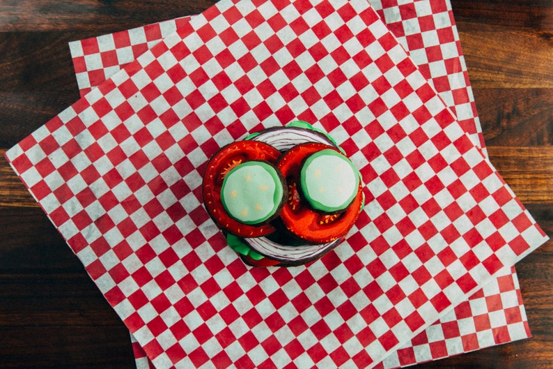 Puede incluir: Un papel a cuadros rojo y blanco con una galleta en capas decorada para parecer una hamburguesa. La hamburguesa de galleta tiene glaseado rojo y verde y un c&iacute;rculo de glaseado blanco en la parte superior.