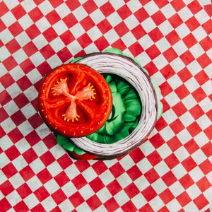 Puede incluir: Una rodaja de tomate rojo, una hoja de lechuga verde y un anillo de cebolla morada est&aacute;n dispuestos sobre un papel a cuadros rojo y blanco. Los alimentos est&aacute;n dispuestos en c&iacute;rculo, asemej&aacute;ndose a una hamburguesa.