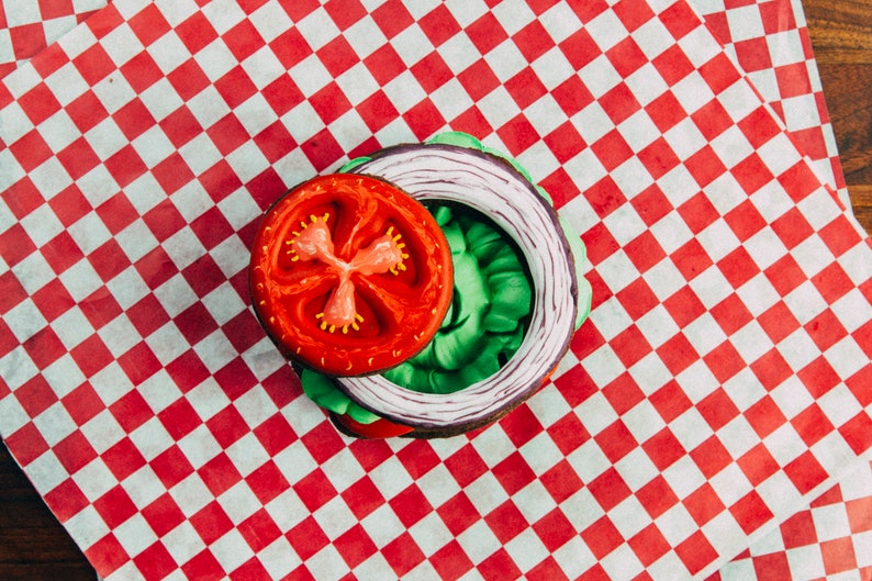 Puede incluir: Una rodaja de tomate rojo, una hoja de lechuga verde y un anillo de cebolla morada est&aacute;n dispuestos sobre un papel a cuadros rojo y blanco. Los alimentos est&aacute;n dispuestos en c&iacute;rculo, asemej&aacute;ndose a una hamburguesa.