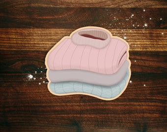 May include: A stack of three knitted sweaters in pink, purple, and blue. The sweaters are folded and stacked on top of each other. The sweaters are on a brown wooden surface.