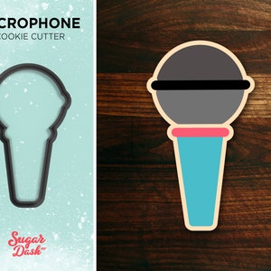 May include: A black cookie cutter in the shape of a microphone. The cookie cutter is on a light blue background with white speckles. The text "MICROPHONE COOKIE CUTTER" is above the cookie cutter. The text "Sugar Dash" is below the cookie cutter.