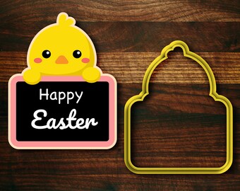 Op de afbeelding: Een geel kuiken met een roze snavel en wangen houdt een roze bord vast met de tekst "Happy Easter" in zwarte letters. Rechts van het kuiken bevindt zich een gele omtrek van een koekjesvormpje in de vorm van een bel.