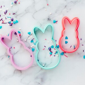 May include: Three bunny-shaped cookie cutters in pink, blue and red, scattered on a white marble surface with colourful sprinkles.