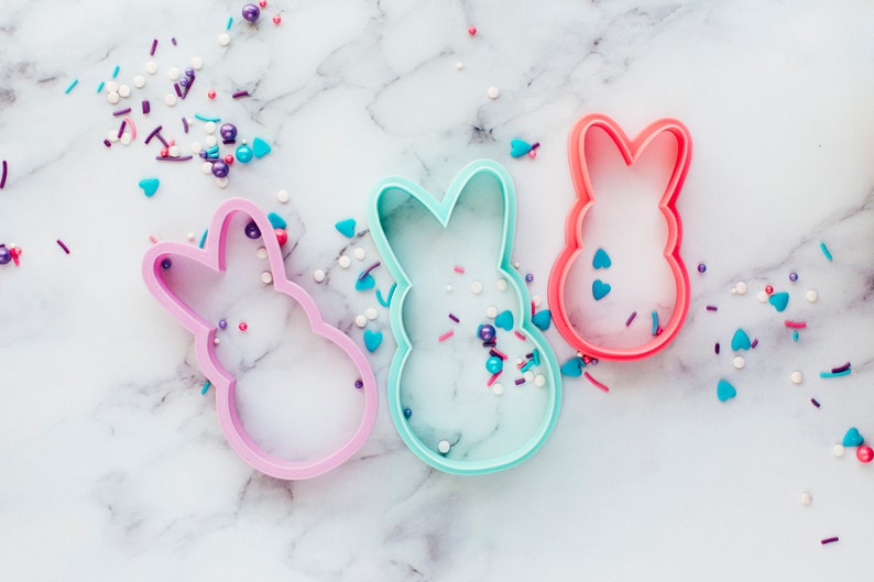 May include: Three bunny-shaped cookie cutters in pink, blue and red, scattered on a white marble surface with colourful sprinkles.