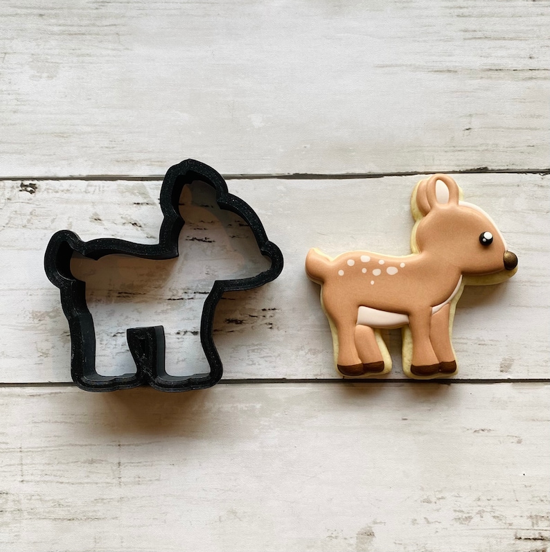 May include: A black plastic cookie cutter shaped like a deer, next to a decorated sugar cookie in the shape of a deer. The cookie is brown with white spots and has a brown nose.