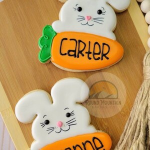 May include: Two decorated sugar cookies shaped like bunnies holding carrots. The carrot on the top cookie has the name "Carter" written on it. The carrot on the bottom cookie has the name "Anne" written on it.