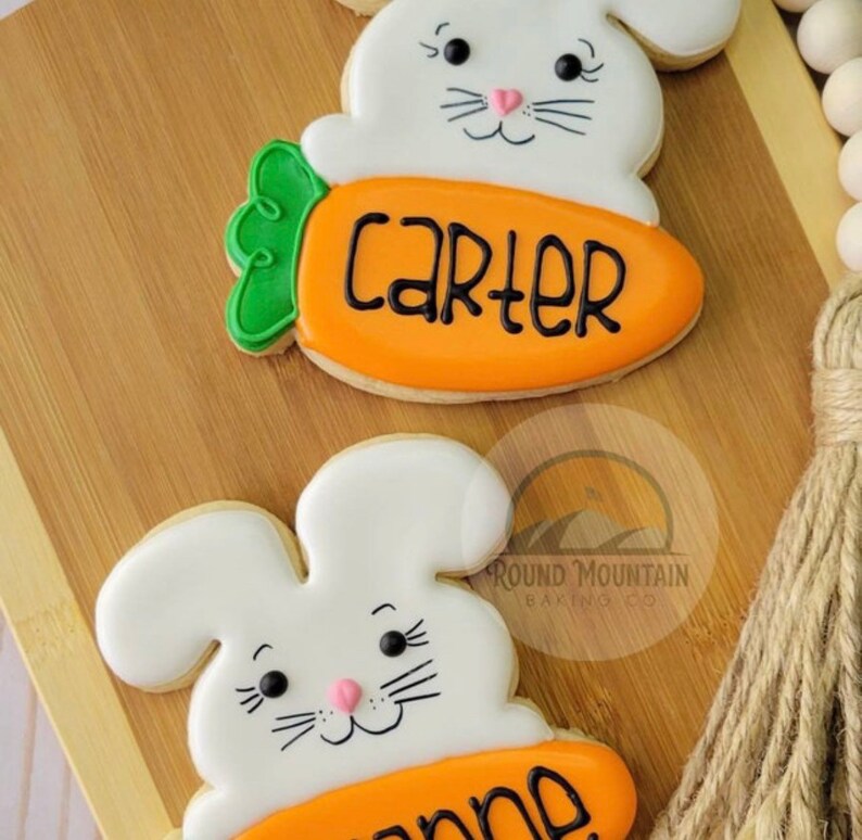 May include: Two decorated sugar cookies shaped like bunnies holding carrots. The carrot on the top cookie has the name "Carter" written on it. The carrot on the bottom cookie has the name "Anne" written on it.