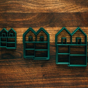 May include: Three green plastic cookie cutters shaped like houses with different sizes. The largest cutter has two windows and a door, the middle cutter has two windows, and the smallest cutter has two windows.
