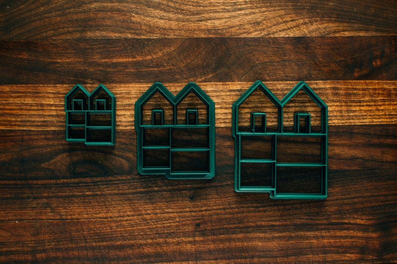 May include: Three green plastic cookie cutters shaped like houses with different sizes. The largest cutter has two windows and a door, the middle cutter has two windows, and the smallest cutter has two windows.