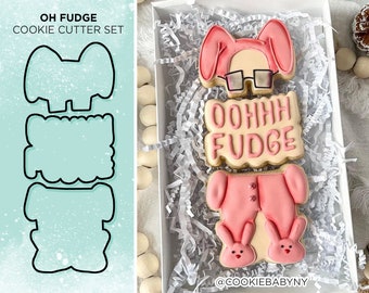 May include: A set of four cookie cutters shaped like a bunny wearing pajamas, a bunny head, a banner that says "OOOHHH FUDGE", and a pair of bunny slippers. The cookie cutters are made of metal and have a silver finish.