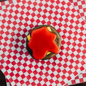 Puede incluir: Una galleta decorada en rojo, blanco y negro con forma de hamburguesa. La galleta est&aacute; sobre un papel de cuadros rojos y blancos.
