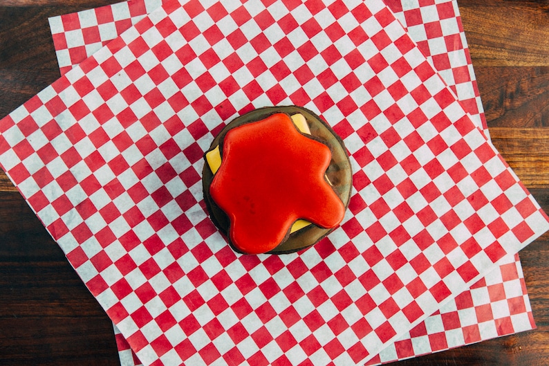 Puede incluir: Una galleta decorada en rojo, blanco y negro con forma de hamburguesa. La galleta est&aacute; sobre un papel de cuadros rojos y blancos.