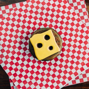 Puede incluir: Una dona de chocolate con glaseado amarillo y tres agujeros, que se asemeja a una rebanada de queso, sobre un papel a cuadros rojo y blanco.