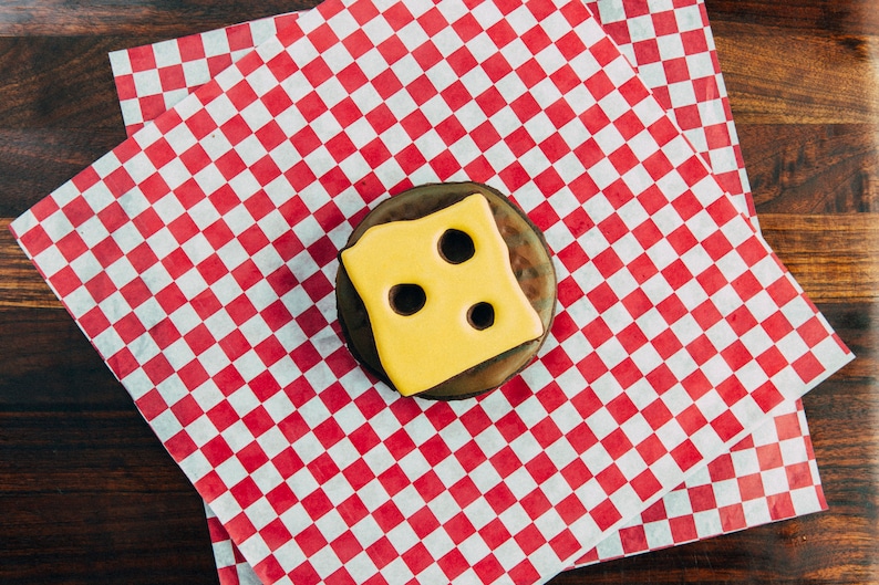 Puede incluir: Una dona de chocolate con glaseado amarillo y tres agujeros, que se asemeja a una rebanada de queso, sobre un papel a cuadros rojo y blanco.