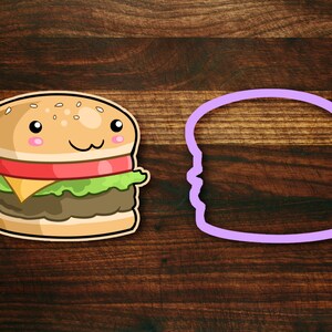 May include: A cartoon burger cookie cutter with a smiling face and a purple outline. The cutter is on a wooden surface next to a cartoon burger with a sesame seed bun, lettuce, tomato, cheese, and a beef patty.
