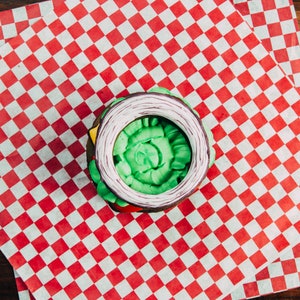 Puede incluir: Una dona verde, blanca y morada decorada con forma de hamburguesa. La dona est&aacute; sobre un papel de cuadros rojos y blancos.