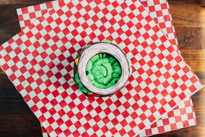 Puede incluir: Una dona verde, blanca y morada decorada con forma de hamburguesa. La dona est&aacute; sobre un papel de cuadros rojos y blancos.