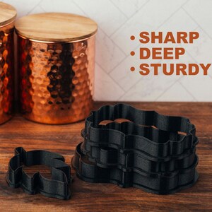 May include: A set of four black cookie cutters, featuring a wavy, ornate design. The cutters are stacked on top of each other. The text "SHARP", "DEEP", and "STURDY" is written in orange on a white background. The text "Sugar Dash co." is written in white on a black background.