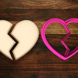 May include: A broken heart cookie cutter in pink plastic, next to a white broken heart on a brown wooden surface.