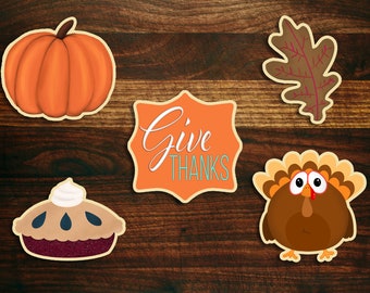 May include: Five Thanksgiving-themed decorations on a wood background. The decorations include a pumpkin, a pie, a turkey, a leaf, and a sign that says "Give Thanks".