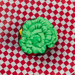 Puede incluir: Una galleta glaseada verde y roja con forma de hamburguesa sobre un papel de cuadros rojos y blancos.