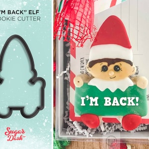 May include: A black cookie cutter in the shape of an elf, with the text "I'M BACK" ELF COOKIE CUTTER. Next to it is a decorated elf cookie holding a green sign that says "I'M BACK!" in white letters. The cookie has a red hat and feet.
