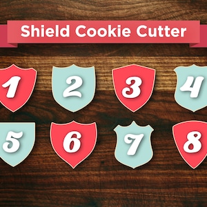 May include: Eight shield-shaped cookie cutters, numbered 1 through 8, in alternating colors of red and light blue. The cookie cutters are arranged in two rows of four on a wooden surface.