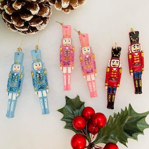 May include: A selection of festive nutcracker earrings in blue, pink, and red. The earrings feature gold detailing and glitter, evoking a holiday theme. The image also includes a pine cone, holly, and red berries, enhancing the seasonal aesthetic.