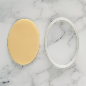 May include: A white oval cookie cutter and a round piece of yellow fondant on a white marble surface.