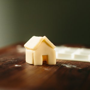 May include: A small, white, house-shaped cookie cutter made of silicone sits on a wooden surface.