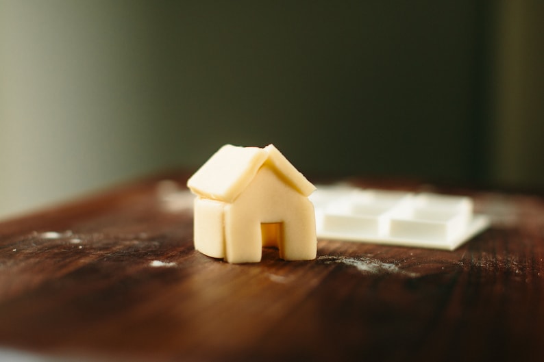 May include: A small, white, house-shaped cookie cutter made of silicone sits on a wooden surface.
