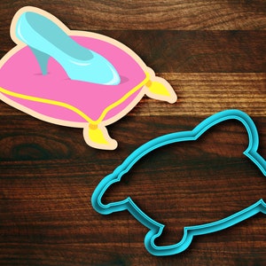 May include: A blue glass slipper on a pink pillow with yellow trim. The pillow is on a wooden surface. There is a blue plastic cookie cutter in the shape of a slipper on the right side of the image.