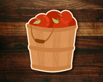 May include: A wooden bucket filled with red apples. The bucket has a brown handle and is sitting on a brown wooden surface.