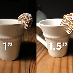 May include: Two white ceramic mugs with gingerbread house cookie stirrers. The mug on the left has a 1" gingerbread house stirrer and the mug on the right has a 1.5" gingerbread house stirrer.