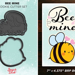 May include: A set of two cookie cutters, one shaped like a bee and the other shaped like the words "Bee Mine". The cutters are black and are shown on a light blue background with a glittery texture. The text "BEE MINE COOKIE CUTTER SET" is at the top of the image. The text "Sugar Dash" is at the bottom of the image. The text "7" x 4.375" BRP BOX" is at the bottom of the image.