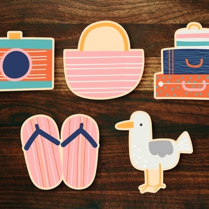 May include: A set of five illustrated stickers featuring a camera, a beach bag, a stack of suitcases, a pair of flip flops, and a seagull. The stickers are all in a cartoon style and have a bright, colorful design.