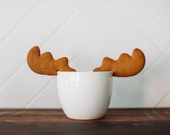 May include: A white ceramic mug with two gingerbread cookies shaped like antlers on top. The mug is sitting on a wooden surface.