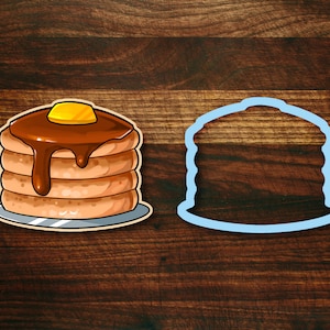 May include: A cartoon illustration of a stack of pancakes with chocolate syrup and a pat of butter on top. A blue outline of the pancakes is shown next to the illustration.