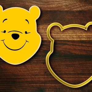 May include: Yellow Winnie the Pooh cookie cutter with a smiling face and closed eyes. The cutter is on a brown wooden surface.