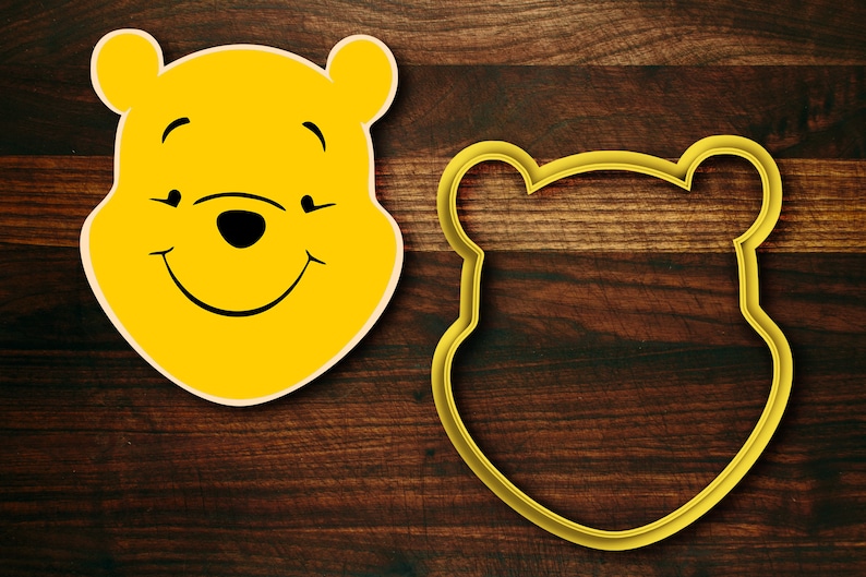 May include: Yellow Winnie the Pooh cookie cutter with a smiling face and closed eyes. The cutter is on a brown wooden surface.